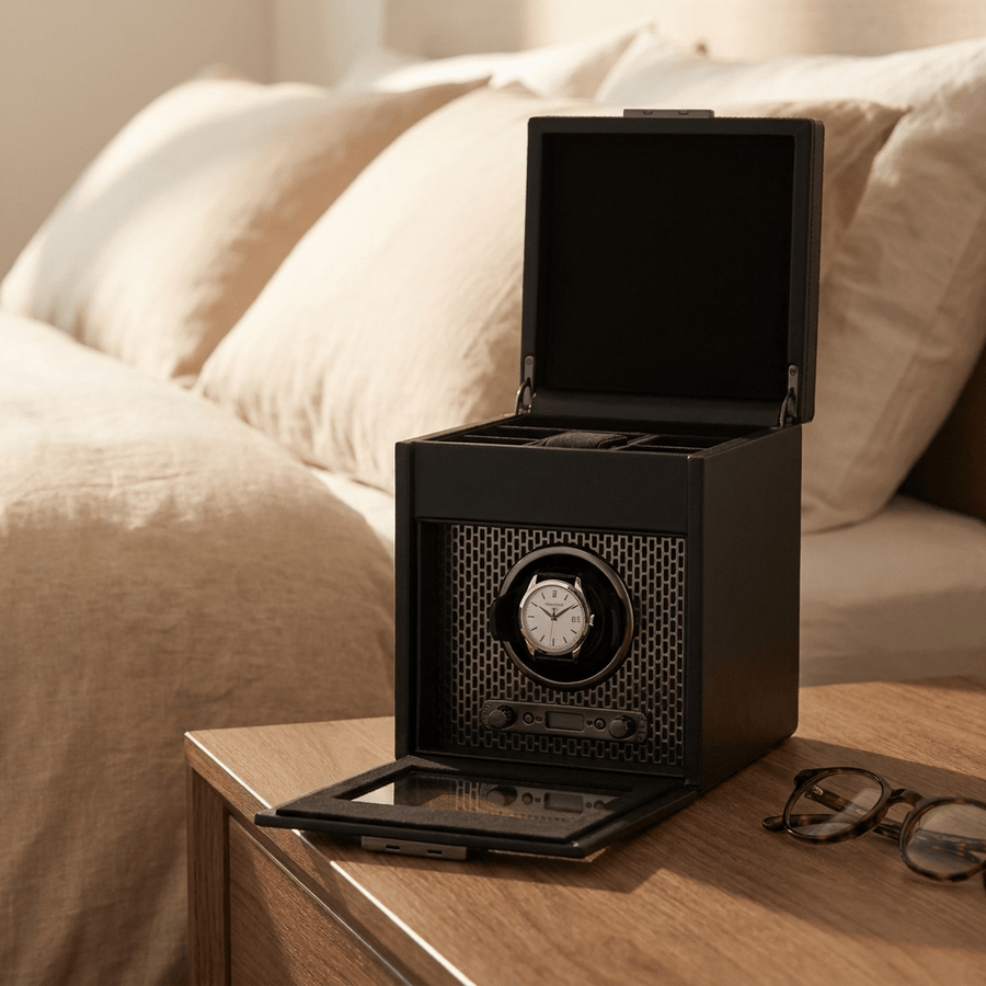 Wolf 1834 Axis watch winder met lade voor 1 horloge in powder coat (mat zwart) afwerking, gevuld met verschillende luxe horloges, op een nachtkastje